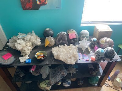 Crystal display table with large quartz clusters, amethyst geodes, and tumbled stones against turquoise wall