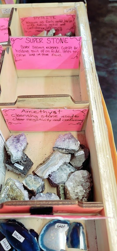Wooden display bins with labeled crystals including pyrite, super stone, amethyst, rose quartz, and clear quartz